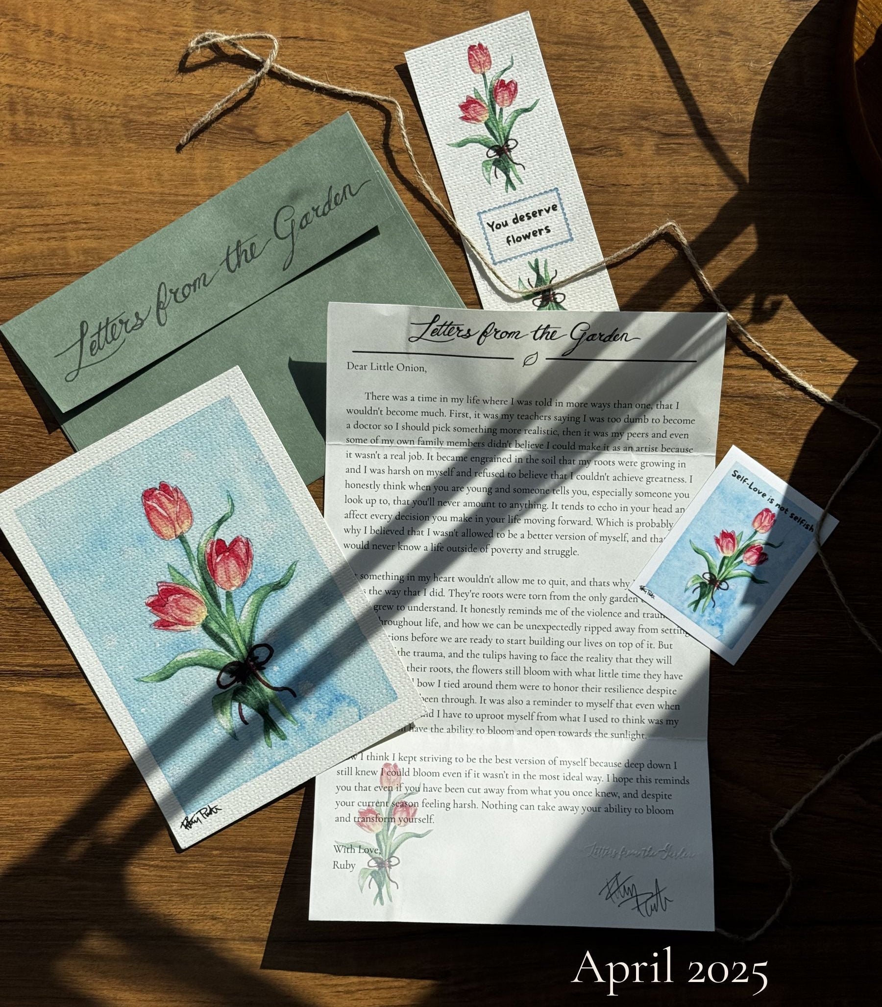 wooden desk with a spread of letter mail contents, showing a letter with black text, watercolor art print of red tulips on a blue background a sticker with the same artwork captioned self love is not selfish and a white bookmark with the red tulips and text saying you deserve flowers
